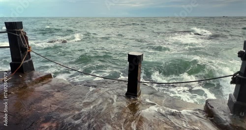 Rough ocean waves crash against a stone pier, sending spray across a black mooring post. ( in slow motion)