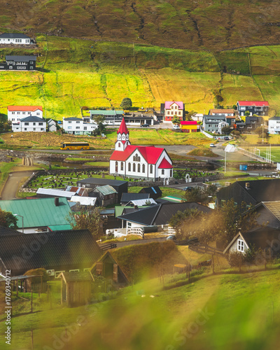 View of a charming village nestled in green hills with a red-roofed church as a focal point, houses scattered across the landscape, Sandavágur, Vágar, Faroe Islands.