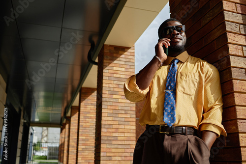Black man wearing sunglasses standing outdoors leaning against brick column talking on smartphone, appearing confident and professional, young adult in business attire