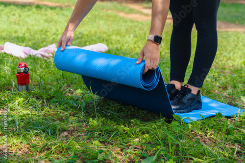 Active woman wearing sportswear rolling blue yoga mat after workout outdoors in green park, concept of fitness, wellness, relaxation and healthy lifestyle routine in nature after physical exercise