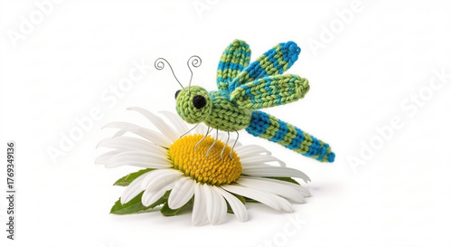 A small, green and blue knitted dragonfly with wire antennae perches on the yellow center of a white daisy flower.