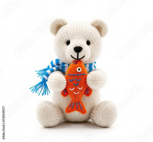 A small, cream-colored knitted bear wearing a blue and white striped scarf sits while holding an orange knitted fish toy.