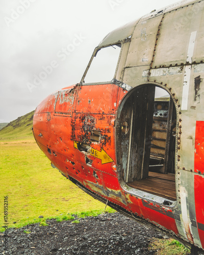 View of the weathered, red-nosed DC-3 airplane wreck, a stark contrast against the vibrant green fields, under a cloudy sky, Eyvindarholt, Rangárping eystra, Iceland.