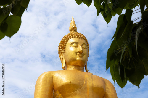 Golden Buddha statue at Wat Muang temple in Ang Thong, Thailand, beautifully framed by Bodhi tree leaves under a bright blue sky, representing peace, faith, and Thai spirituality.