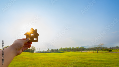 Hand holding wooden house model against sunlight in green field, symbolizing eco-friendly housing, real estate investment, sustainable living, or future home development concept.