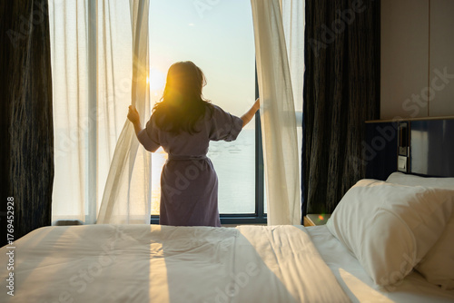 Healthy sleep, Recreation and Relaxation Concept. Happy middle aged woman waking up in bedroom and opens the curtains to let the Soft morning sunlight streaming through window. Greeting new day.