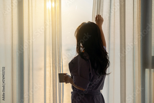 Thoughtful middle aged woman opening curtain for beautiful sunshine, thinking about future in cozy apartment in moment of quiet introspection while drinking morning coffee by the window in bedroom.