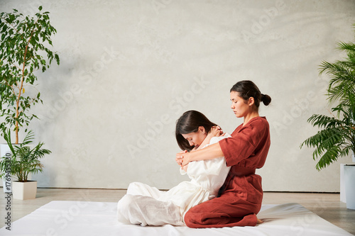 Masseuse making thai yoga massage. Woman therapist doing traditional massage treatment, stretching female patient's body. Client sitting on mat, enjoying hand massage in calm atmosphere.