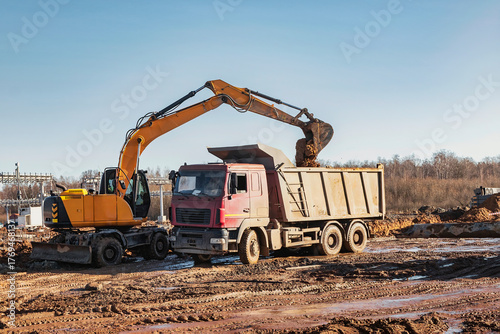 A wheeled excavator loads a dump truck with soil and sand. An excavator with a high-raised bucket against a cloudy sky View from the trench. Removal of soil from a construction site or quarry.