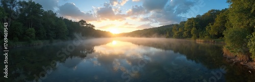 Sunrise mist drifts over calm lake water reflecting sky clouds and trees. Green forested hills border serene river at dawn. Gentle light breaks through mist creating peaceful morning atmosphere.