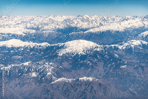 Aerial view of snow-capped peaks pierce the sky, a rugged landscape etched in white and grey under a clear blue horizon, somewhere over the mountains.
