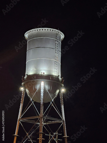 Nighttime architecture image of a tall steel water tower illuminated by spotlights, taken from below the structure while looking up at it.