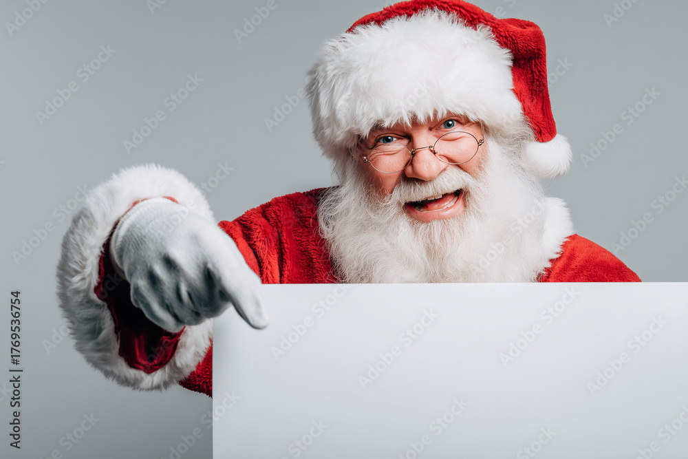 Naklejka premium Joyful Santa dressed in classic red pointing toward a blank signboard, expressing holiday cheer