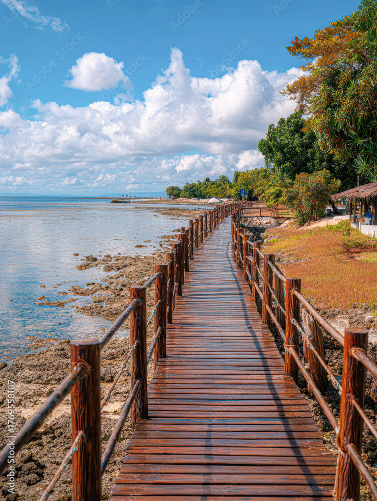 Naklejka premium A serene wooden walkway stretches along the shoreline, bordered by lush greenery and vibrant clouds, leading to a peaceful coastal view.