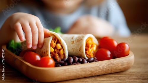 A child is eating a burrito with tomatoes and beans. The child is holding the burrito with one hand and a tomato with the other