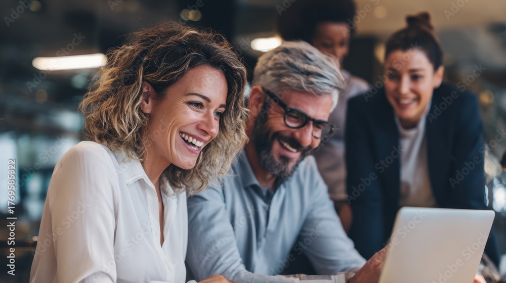 © Formoney - Colleagues smile and engage around a laptop in a bright office sharing ideas and laughter together.