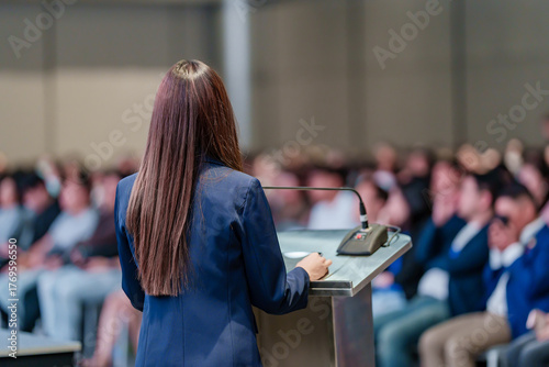 Rear view of woman speaker giving an inspiring talk or leading a workshop for young professionals and adult training participants in a large conference room.