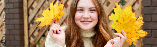 Beautiful smiling redhead teen girl with maple yellow leaves over wooden background. Autumn time. Fall season. knitted sweater