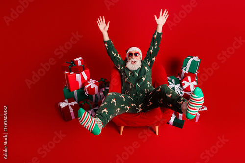 Cheerful Santa in festive pajama suit sits in red chair surrounded by gifts with striped socks celebrating christmas eve at home