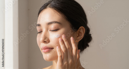 An Asian woman applying skincare cream to her face with eyes closed, enjoying the sunlight.