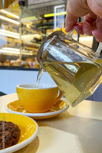 A woman holds teapot and pours tea into yellow cup. Coffee shop interior. Blurred background. Vertical. Close-up
