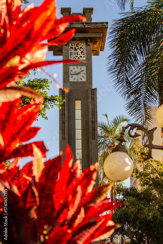 Detalhe da torre do relógio em estilo Art Deco na  Avenida Goiás em Goiânia, emoldurado por plantas de folhas vermelhas em um dia claro e ensolado.