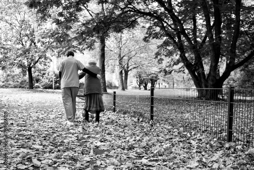 Elderly woman taking a walk through the park in autumn with help of walker and caretaker aid