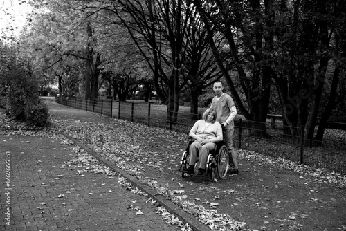 Happy elderly woman with grey hair sitting in wheelchair during walk in park with help of caretaker in blue uniform