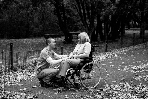 Happy elderly woman with grey hair sitting in wheelchair during walk in park with help of caretaker in blue uniform