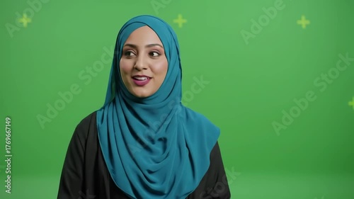 Woman in teal hijab smiles and speaks against a green screen background.