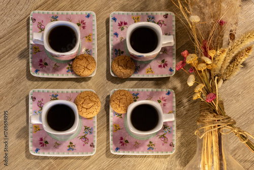 Four cups of coffee with cookies on floral saucers over a wooden table beside a bouquet of dried flowers, representing the social and cultural coffee tradition in Brazil.