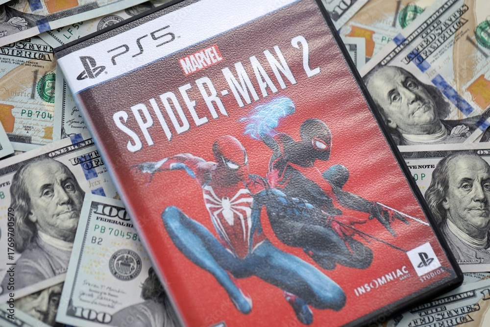 Naklejka premium KYIV, UKRAINE - SEPTEMBER 8, 2025 Marvels Spider-Man 2 videogame lies on big amount of hundred dollar bills close up