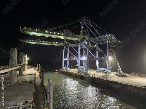 Night container terminal with giant gantry cranes, tugboat, and fishing vessels reflecting lights on water.