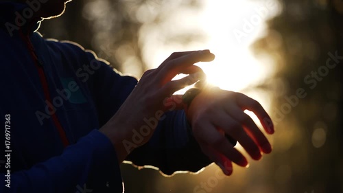 Close-up of a silhouetted athlete using a smart watch to track performance during a training run in a park. The morning sun creates a beautiful lens flare, illuminating the hands