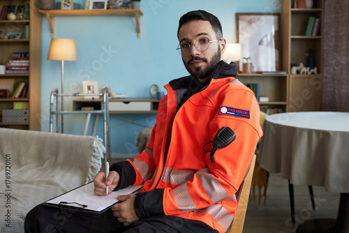 Portrait of young Middle Eastern male paramedic with short beard and glasses sitting indoors wearing emergency responder jacket holding pen and clipboard looking at camera, during patient call