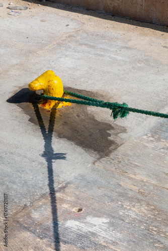 A bright yellow mooring bollard secures a green rope on a weathered concrete dock, casting long shadows under strong sunlight.