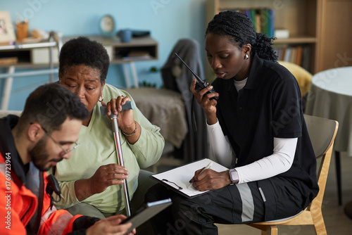 Middle aged Black woman holding cane sitting next to young female nurse using walkie talkie and clipboard with male paramedic showing information on tablet, discussing emergency hospitalization plan