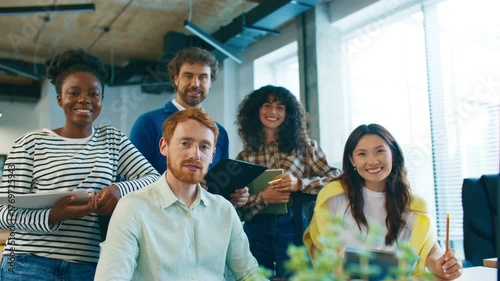 Group of coworkers pausing discussion and looking directly at camera with focused expressions. People standing and sitting in modern office environment holding tablets and notebooks.