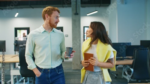 Smiling coworkers spending break together. Male with beard holding coffee cup and woman carrying notebook talking cheerfully. Sharing friendly moment between tasks. Standing near desks in office area.