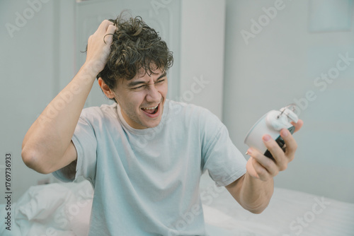 Cuadro en lienzo Young man looking frustrated while holding alarm clock and scratching his head in the morning