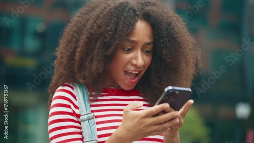 African American female with curly hair wearing striped shirt and denim overalls looking at smartphone with wide smile after receiving good news. Messaging online with friends outdoors near office.