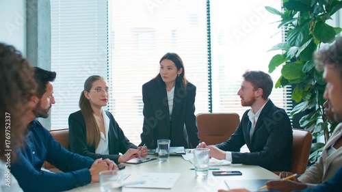 Business team sitting around table listening to standing woman presenting issue. Everyone focused and thoughtful, facing difficult decision. Serious discussion scene in meeting room.