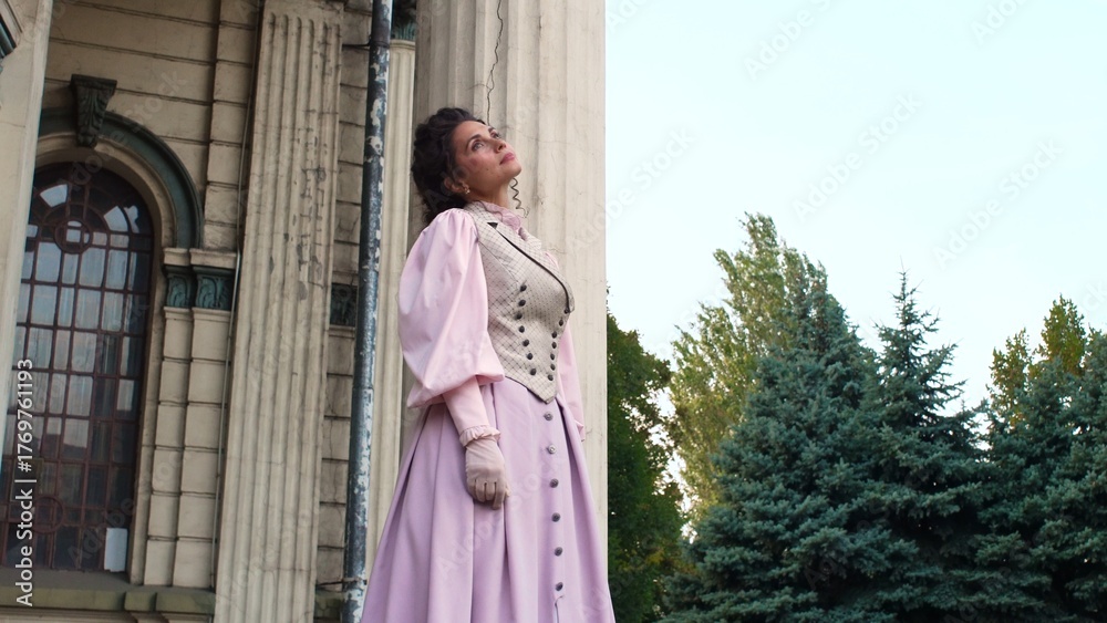 Fototapeta premium An elegant woman in a pastel historical dress stands next to a weathered column of an old building, with a dreamy, nostalgic expression on her face.