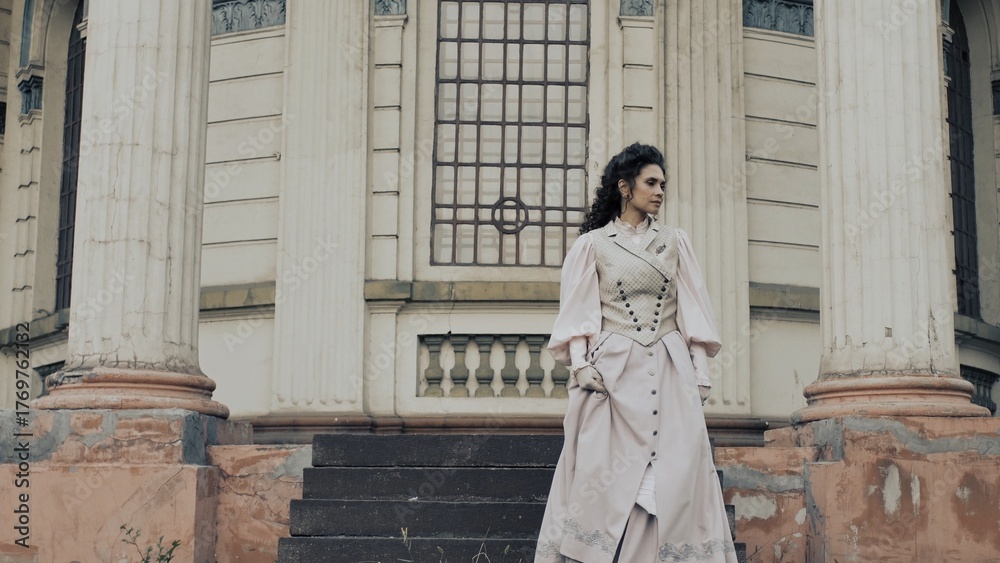 Fototapeta premium An elegant woman in a pastel historical dress stands next to a weathered column of an old building, with a dreamy, nostalgic expression on her face.