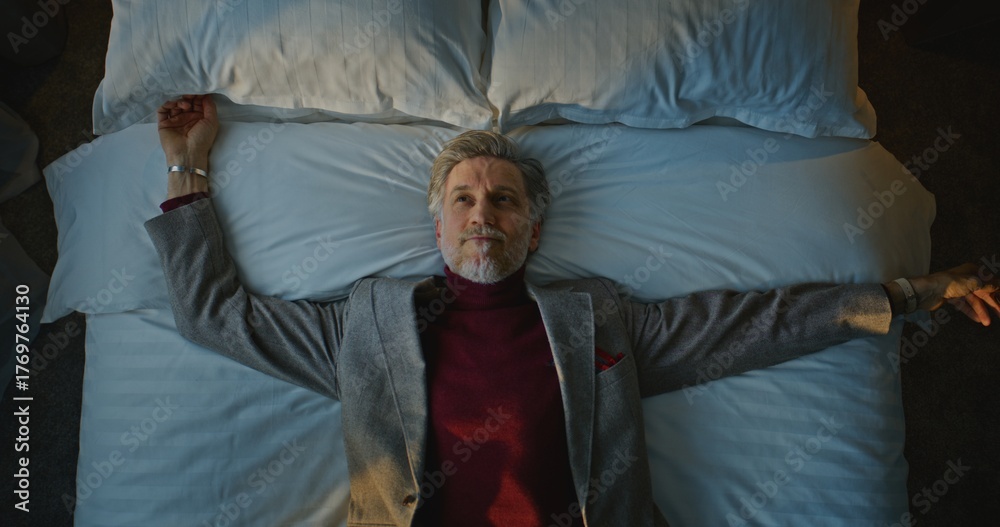 Fototapeta premium Aged Man in Suit Happily Falls Down on White Bed in Cozy Hotel Room. Tired From Traveling on Business Trip, Man Rests on Bed. Concept of Life in Hotel, Traveler Fatigue, Business Trip. Top View.