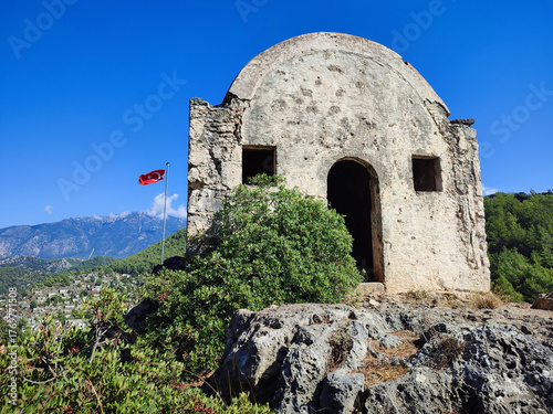 An ancient sanctuary on a rocky plateau against the ghost town of Kayaköy and flag of Turkey