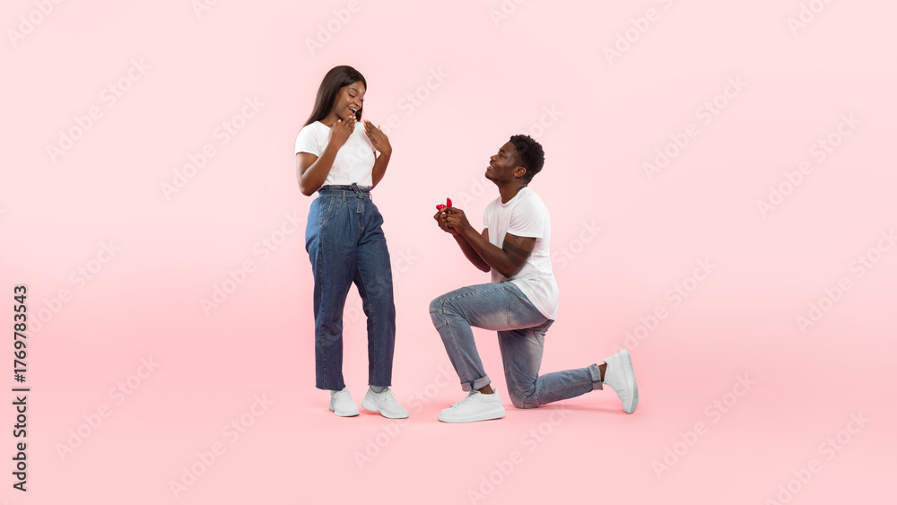 Naklejka premium A joyful black man kneels and presents an engagement ring to his thrilled girlfriend, asking her to marry him. They are in front of a soft pink background, capturing a beautiful moment of love.