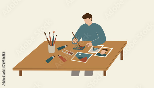 Man holding a paintbrush and painting with watercolors on a table, surrounded by art supplies and framed illustrations, emphasizing creativity and hobby