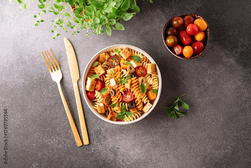 Warm salad with colorful full grain fusilli pasta, olives, fried tofu, cherry tomatoes and aromatic oregano leaves in ceramic bowl on black stone table top view. Healthy vegan food concept.