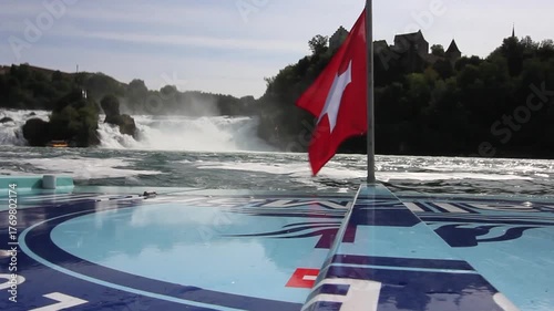 A boat-level perspective of the Rhine Falls, featuring the Swiss flag waving in the foreground and Laufen Castle visible atop the cliff overlooking Europe's largest waterfall